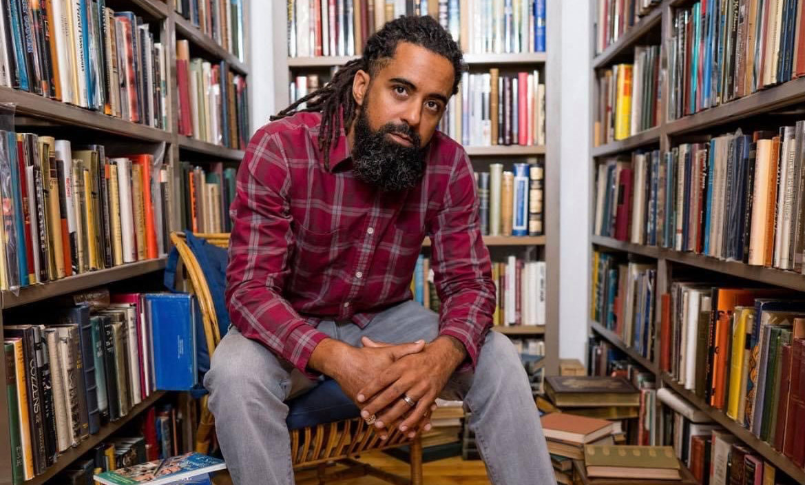 Jesse Kelley sits in front of shelves of books with books stacked on the floor