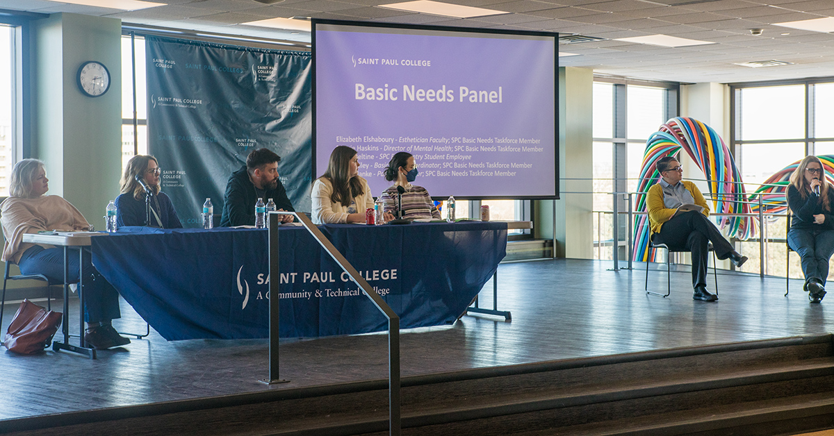 Several speakers at a table in front of a projection that reads basic needs panel